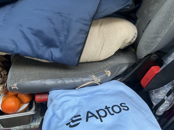 A cluttered vehicle trunk or storage area containing blue Aptos-branded bags and jackets alongside oranges and various supplies.
