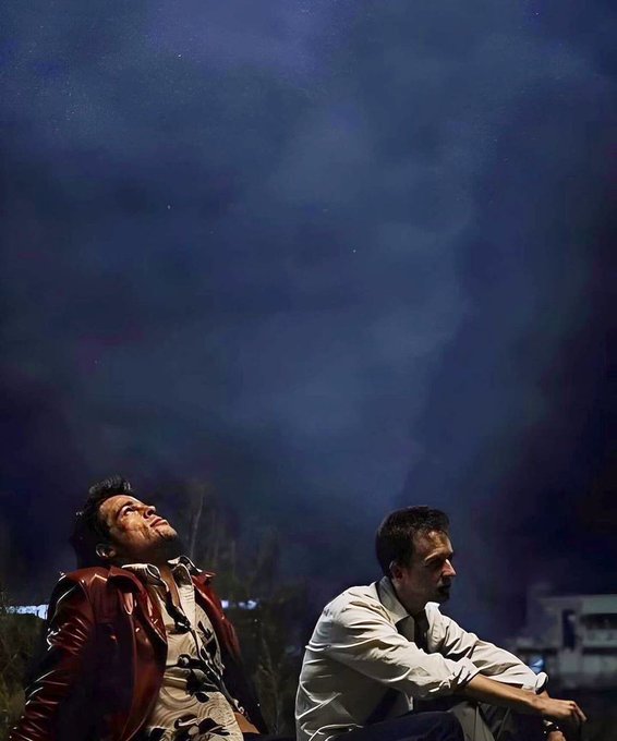 Two men gaze upward at a night sky dominated by a large, softly glowing moon, capturing a quiet moment of shared wonder under the celestial display.