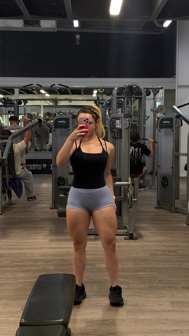A woman takes a mirror selfie at the gym wearing a black tank top and light blue shorts, with "KAESUN ARMY" visible on equipment behind her, showcasing her leg day progress as part of her "Today's pump!" routine.