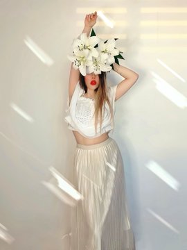 A woman in a white crop top and pleated skirt holds a bouquet of white flowers over her face, with light streaming across the plain background.