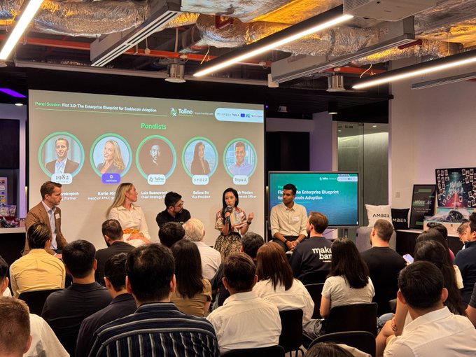 The image shows a group of people sitting around a table with a projector screen behind them. The audience appears engaged, possibly listening to a presentation or panel discussion about the future of enterprise stablecoins. The room has a modern design, featuring an open floor plan that allows for easy interaction among attendees.