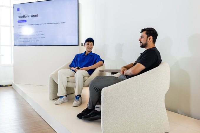 Two men sit in upholstered armchairs on a small white platform during a discussion; a screen reads “Base Memo Summit.”