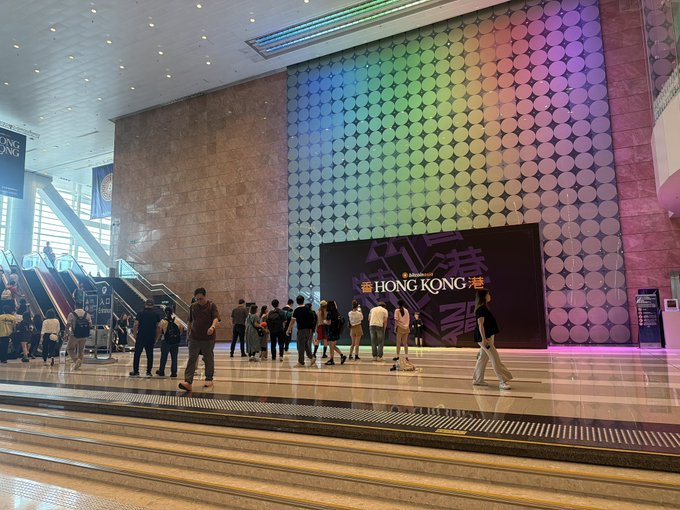 Interior shot of Hong Kong International Airport featuring a large illuminated wall display with rainbow lighting and 'HONG KONG' signage, with travelers and escalators visible in the foreground.