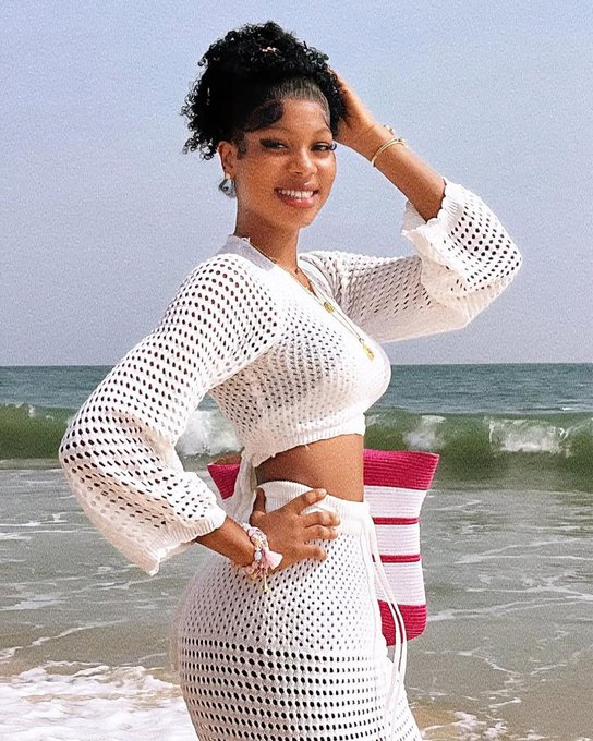 The image features a woman standing at the edge of the ocean wearing a white dress and skirt. She has her hair styled in an updo and is smiling for the camera, conveying a sense of joy and relaxation. The beach setting suggests that she might be enjoying a vacation or spending time with friends and family.