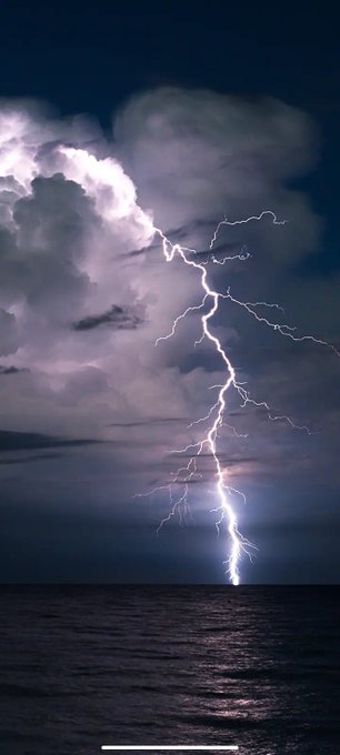 A dramatic nighttime photograph captures a bright lightning bolt striking down from a large, illuminated cloud formation and hitting the surface of the dark ocean.