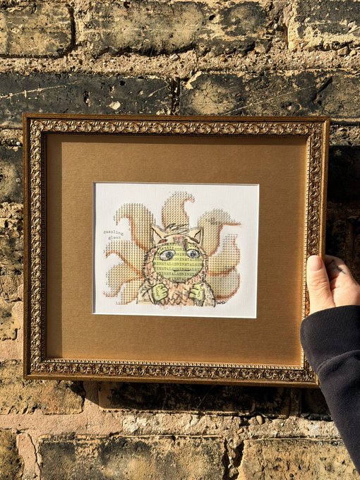 A hand holds a gold ornate frame containing a small cross-stitch artwork of a green cat-like creature with flame-like features, signed 'sparkling'.