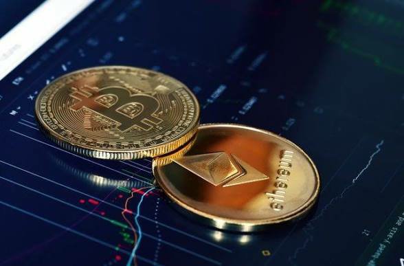 Two physical cryptocurrency coins—a gold Bitcoin and an Ethereum coin—resting on a screen displaying a stock/crypto trading chart with candlestick patterns.
