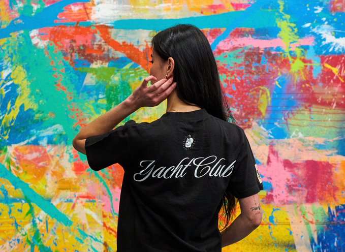 A woman with long dark hair stands facing a vibrant, paint-splattered wall, wearing a black shirt with "Yacht Club" printed in cursive on the back. The image ties into the BAYC x King Saladeen Anchor Drop by showcasing the "Yacht Club" branding associated with the Bored Ape Yacht Club collaboration, where King Saladeen's art style complements the colorful, expressive aesthetic of the collection.