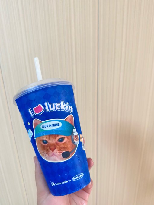 A hand holding a blue Luckin Coffee cup featuring the brand's signature orange cat mascot wearing a "LUCK IN HAND" headband, with a white straw.