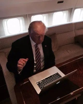 An older man in a suit and striped tie sitting inside what appears to be an aircraft cabin, working on a silver laptop on a wooden table.