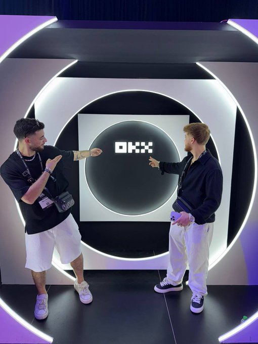 Two men pose and gesture beneath a glowing circular display featuring the OKX logo. The post’s “GM” likely signals a friendly “good morning” message.