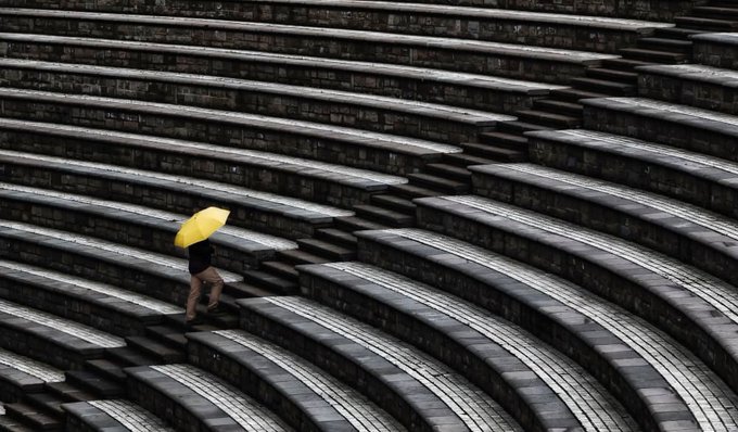 A lone figure with a bright yellow umbrella walks across the curved stone steps of an amphitheater in a striking black and white composition.