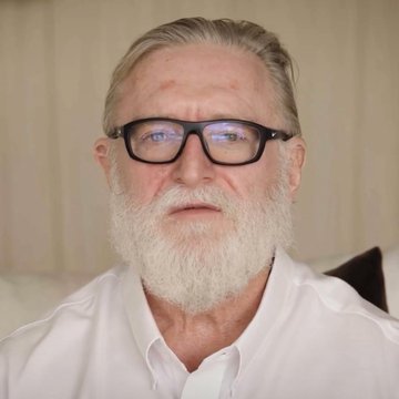 A close-up portrait of an elderly man with a white beard wearing black-rimmed glasses and a white shirt.