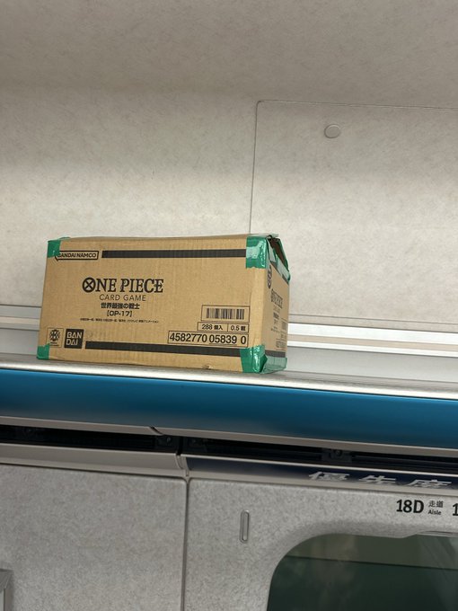 A cardboard box labeled 'ONE PIECE CARD GAME' (OP-17) by BANDAI resting on a shelf inside what appears to be a train car.