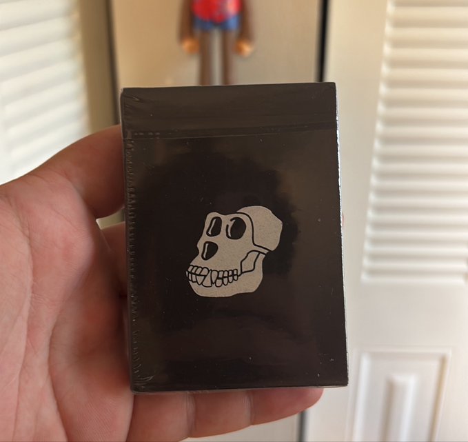 A hand holds up a small black sealed package featuring a white cartoon skull illustration, with a small figure visible in the background.