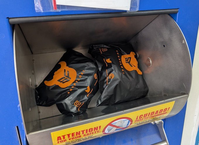 Two black delivery bags with orange Rockauto.com logos sitting inside a metal drop box/receptacle mounted on a blue surface, with a yellow safety warning label reading 'ATTENTION! FOR YOUR SAFETY' and '¡CUIDADO!' visible at the bottom.