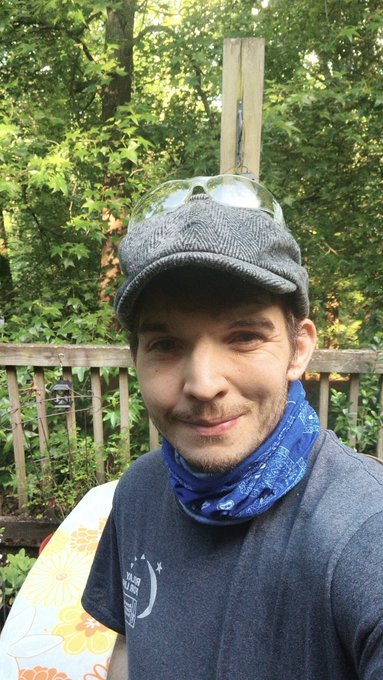 Andy Greensmoke , a man with a blue bandana around his neck, is posing for a photo while wearing a gray cap and sunglasses. The background features trees and a wooden fence, creating an outdoor setting. The caption reads "Who am I... My name is Andy Greensmoke @andygreensmoke_ 2 years clean after discovering crypto. My first buys were $VVV , $Virtuals, and $B3 (two out of three not bad) @AskVenice literally saved my life".