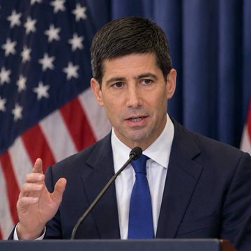 A man in a dark suit and blue tie speaks at a podium with an American flag in the background, gesturing with his hand as he addresses the audience. The accompanying post discusses Fed Chair Kevin Warsh's comments on interest rates, suggesting the image likely depicts him during a press conference or official statement.