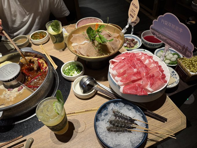 A table full of food at a Chinese hot pot restaurant, featuring thinly sliced raw meat, tripe, spicy hot pot broth, ice with skewers, and various dipping sauces and garnishes.