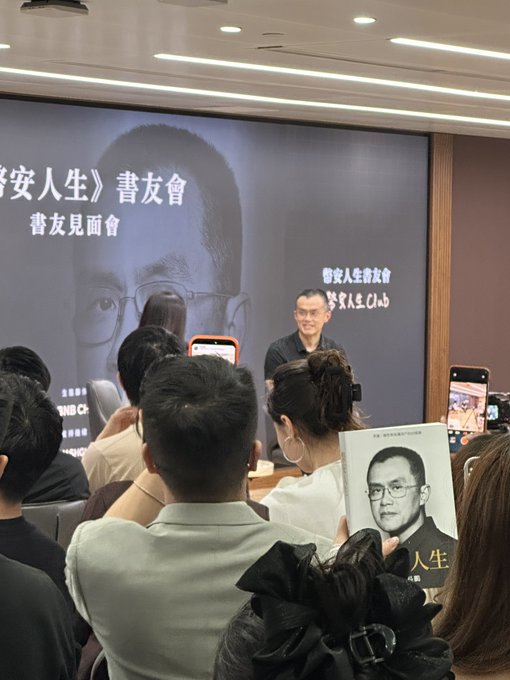 A book launch event titled "幣安人生" (Binance Life) with a "書友見面會" (Book Club Meetup) banner, where the author stands on stage in front of an audience, while attendees hold up phones to record and one person displays a copy of the book featuring the author's portrait on the cover, tying into the #MyBinanceLife (#我的幣安人生) theme.
