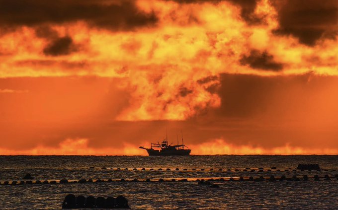 A small fishing boat is silhouetted against a dramatic, fiery orange sunset sky over the ocean, with rows of buoys visible in the foreground water.