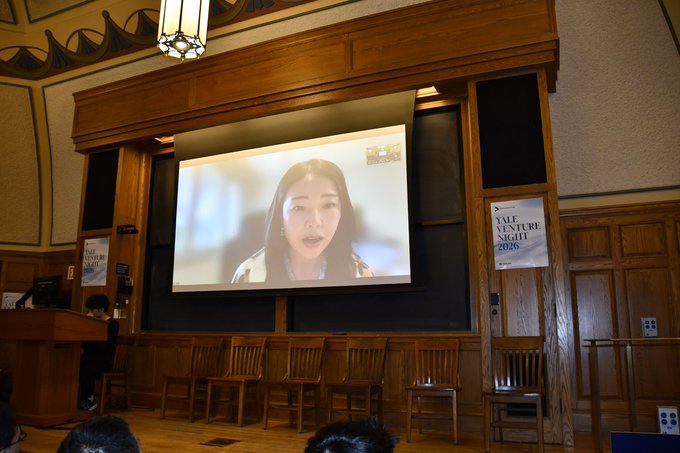 A woman speaks via video on a large projection screen at Yale Venture Night 2026, while a sign bearing the event name hangs on a wooden panel to the right, capturing the speaker portion of the YZi Labs presentation referenced in the post.