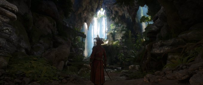 A lone warrior in red robes stands in a lush, overgrown cave looking toward a waterfall cascading down from an opening above.