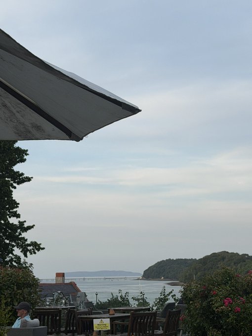 A scenic view from an outdoor cafe under a large white umbrella, looking out over a body of water toward a bridge and tree-covered hills under a cloudy sky.