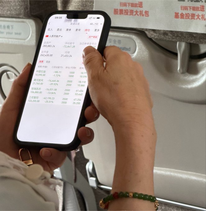 A person holds a smartphone displaying a Chinese stock trading app with significant losses, wearing a green beaded bracelet, sitting on what appears to be public transportation with Chinese text advertisements in the background.