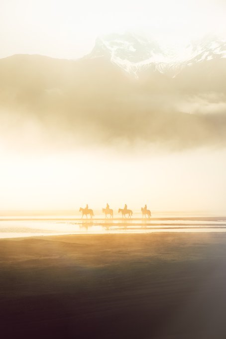 The image shows a group of people riding horses across a sandy beach near a mountain range under a cloudy sky. The photo captures the essence of adventure and exploration as they travel together through this picturesque landscape, with the majestic mountains providing a stunning backdrop for their journey.