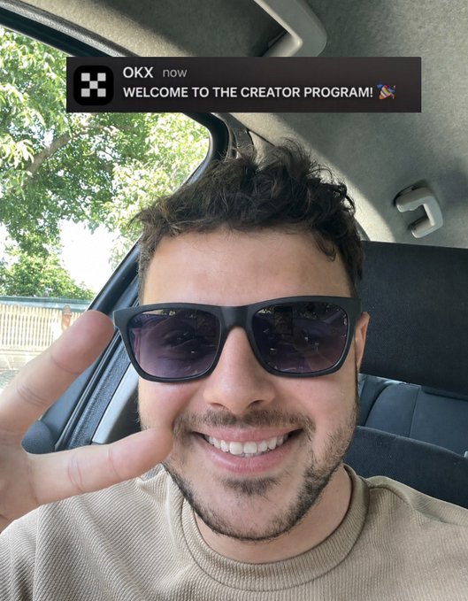 A man wearing sunglasses smiles and flashes a peace sign from inside a car, with a pop-up notification at the top reading "OKX now WELCOME TO THE CREATOR PROGRAM!"—visually celebrating his excitement about joining OKX's creator program.