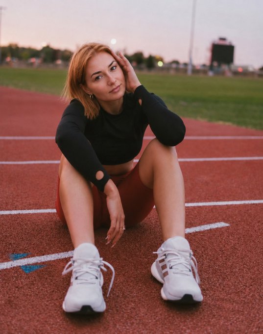 A young blonde woman in a black long-sleeve crop top and maroon shorts sits on a red running track, posing with white sneakers stretched out toward the camera.