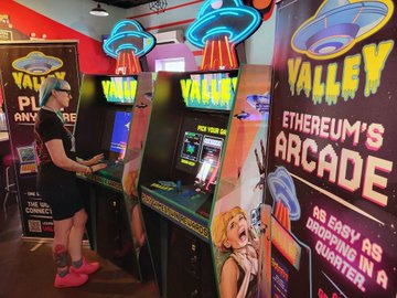 The image features a woman standing next to a booth with two arcade games, one of which has an alien theme. The booth appears to be part of a larger event or gathering related to the Valley Arcade Games. There are four arcade cabinets visible in the scene, and a sign is displayed that reads "Valley Arcade's". A person wearing pink shoes can also be seen near the center of the image.