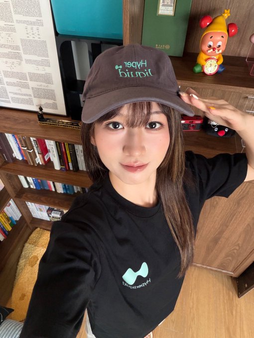 A young woman wearing a black Hyperliquid cap and matching t-shirt takes a selfie in front of a bookshelf with a yellow plush toy visible in the background.