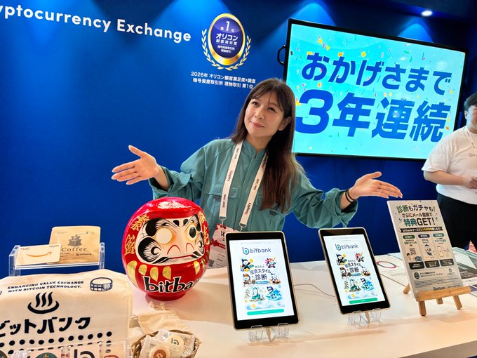 At a bitbank booth, a woman gestures towards tablets displaying an "Investment Style Diagnosis" and a large Daruma doll with the bitbank logo. The backdrop congratulates them on "3 consecutive years," likely referring to a business achievement.