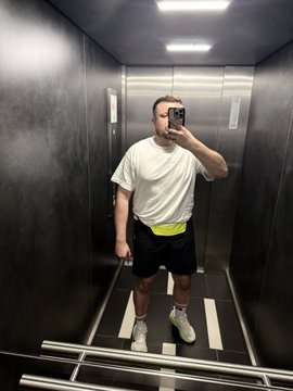 A mirror selfie of a man taking a photo with his smartphone while standing in an elevator, wearing a white t-shirt, black shorts, and white sneakers.
