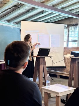 A woman presents in front of a whiteboard and projector screen to a small audience in a rustic classroom, while a participant in the foreground listens attentively alongside a notebook and water bottle—visually capturing the blockchain training session mentioned in the post about the visit to Şirince village with @sinancanw and @RlayHub.