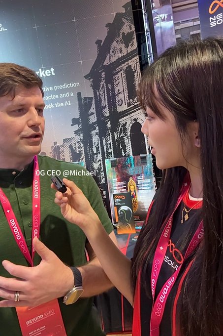 The image features two people standing next to each other, with one person holding a microphone while talking to the other. They are both wearing red lanyards that have "0G" written on them. The background of the image shows an exhibition booth for "0G", and there is a poster hanging on the wall behind the two individuals.