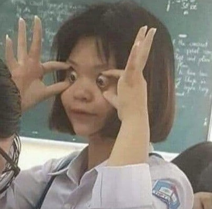 A young student in a school uniform with a shoulder patch poses playfully in front of a chalkboard, making a silly face and holding her hands up around her eyes like binoculars.