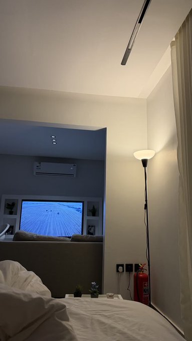 The image shows a bedroom with white walls, a bed covered in white sheets, and a lamp next to the bed. A television screen is mounted on the wall above the bed, displaying an image of a beach or ocean scene. The room also features a couch and a red fire extinguisher placed nearby.