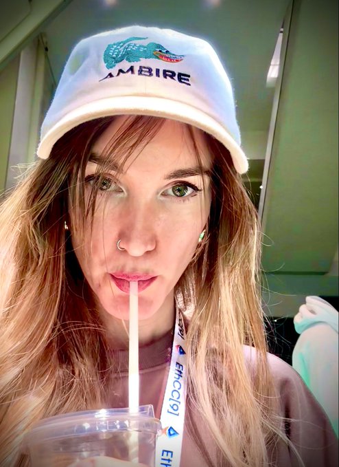 A close-up selfie of a young woman with long blonde hair wearing a white 'AMBIRE' cap with a crocodile logo, drinking from a clear plastic cup with a straw.