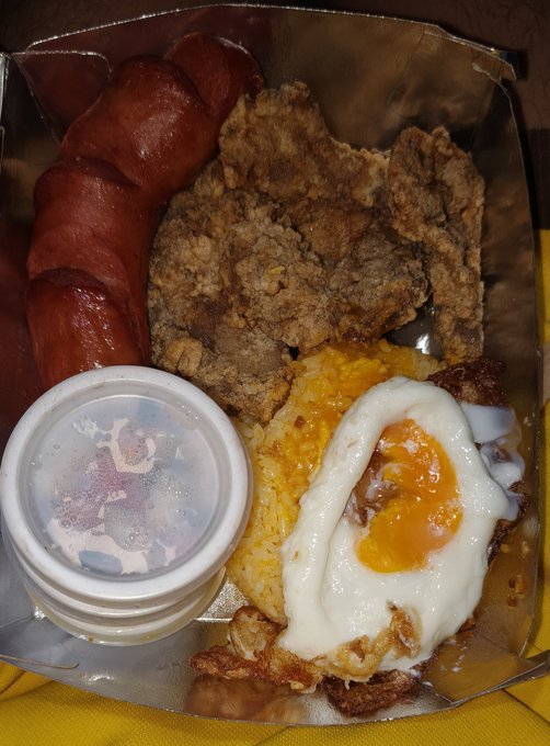 A top-down view of a takeout food container holding a meal of red rice, fried chicken pieces, a fried egg, and a sausage link, alongside a small plastic cup with a lid.