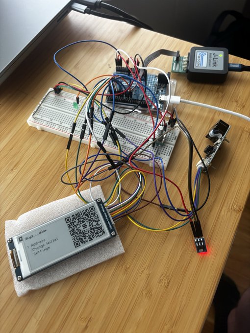 An electronics project on a wooden desk featuring an Arduino board on a breadboard with numerous jumper wires connected to various components, including a small e-paper display showing a QR code and an LED.