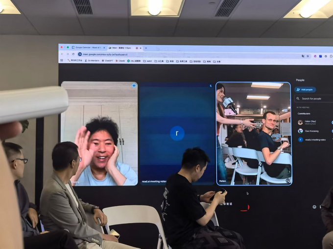 The image shows a group of people sitting around a table, with one person holding up their phone displaying an image or video feed from another location. The people are engaged in conversation while looking at the screen. There are chairs placed around the table where they are seated. A computer monitor is also visible on the wall behind them.