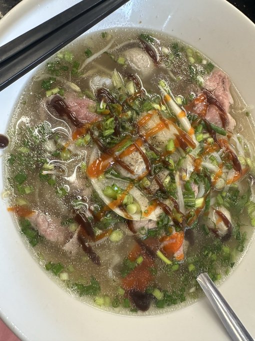 A bowl of Vietnamese pho soup topped with sliced beef, onions, green onions, and drizzled with sriracha and hoisin sauce.