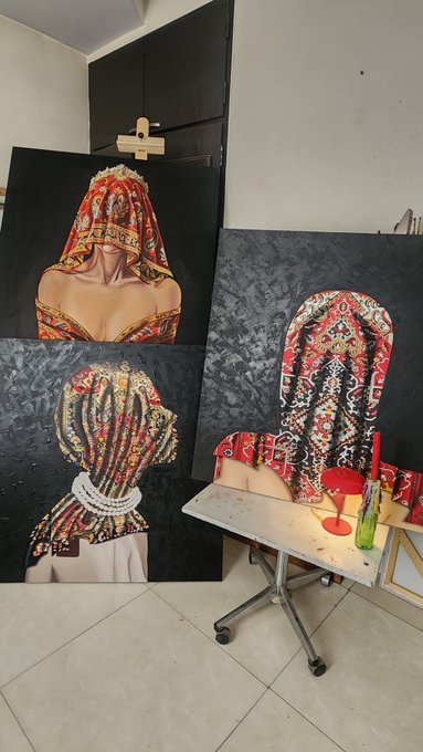 Three large paintings of women wearing ornate, traditional embroidered headpieces and veils are displayed in an art studio next to a rolling cart with paint supplies.