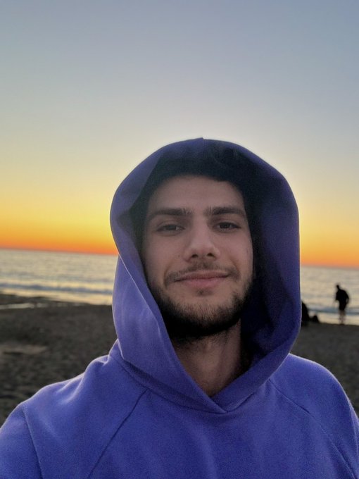The image features a man wearing a blue hoodie, standing at the edge of a beach with the ocean in the background. The sunset casts a warm glow over the scene, creating a serene atmosphere. In the bottom right corner of the photo, there is text that reads "This is epic! See you SOL maxis soon in Istanbul". This message seems to be related to an upcoming event or trip involving the man and his friends.