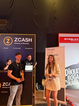 The image shows a group of people standing next to a booth at an event. There are three individuals present - two men and one woman. The man in the foreground is holding a laptop, while the other two people appear to be posing for a photo or attending to their own activities. A banner with the word "ZECASH" written on it can also be seen in the scene.