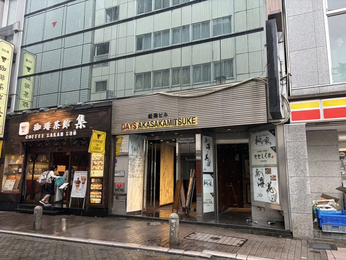 A storefront on a Tokyo street houses "DAYS AKASAKITSUKE" on the ground level alongside "COFFEE SAKAN SOU," with the event taking place on the second floor above these shops. A person in a white shirt is seen near the entrance of the coffee shop.