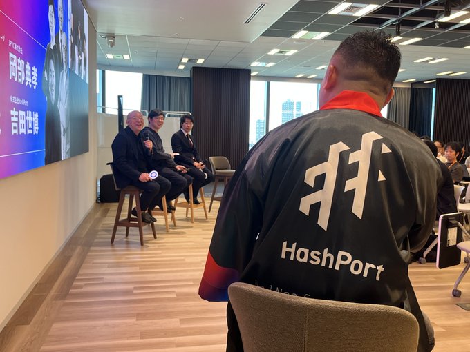 A person wearing a black jacket with 'HashPort' logo and Japanese character sits in the foreground at what appears to be a panel event with speakers on stage.